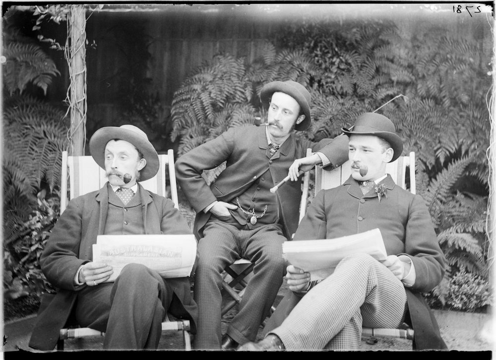 Three men sitting in deck chairs, smoking pipes and reading papers by State Library of Victoria